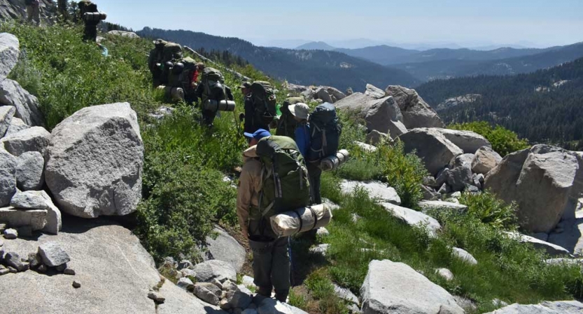 A group of backpackers hike through an area with green shrubs and large boulders. 